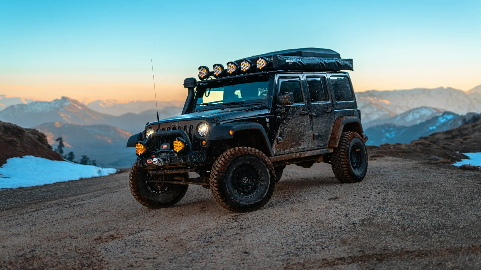 Jeep Wrangler with roof lights on a mountain dirt road at dusk
